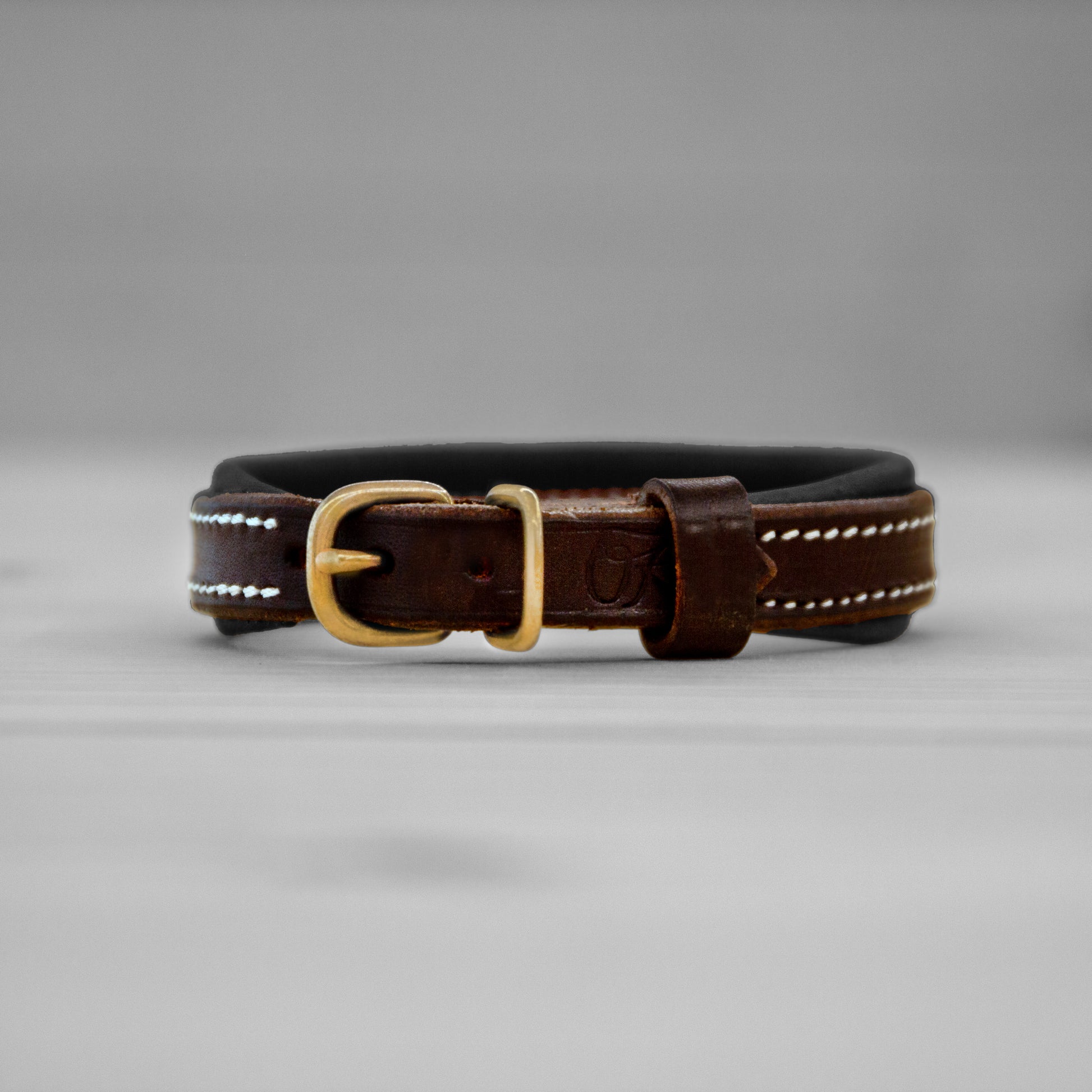Collier en cuir brun avec doublure noire et boucle en laiton pose debout sur un fond gris