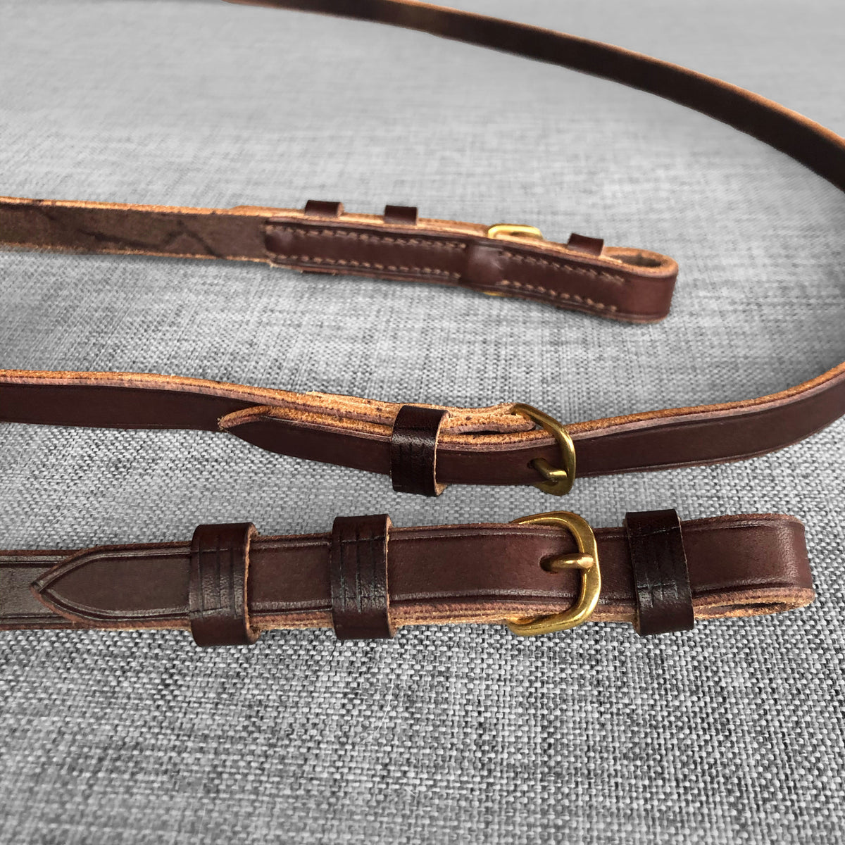 Deux renes en cuir brun avec boucles dorees, empilees sur un fond gris