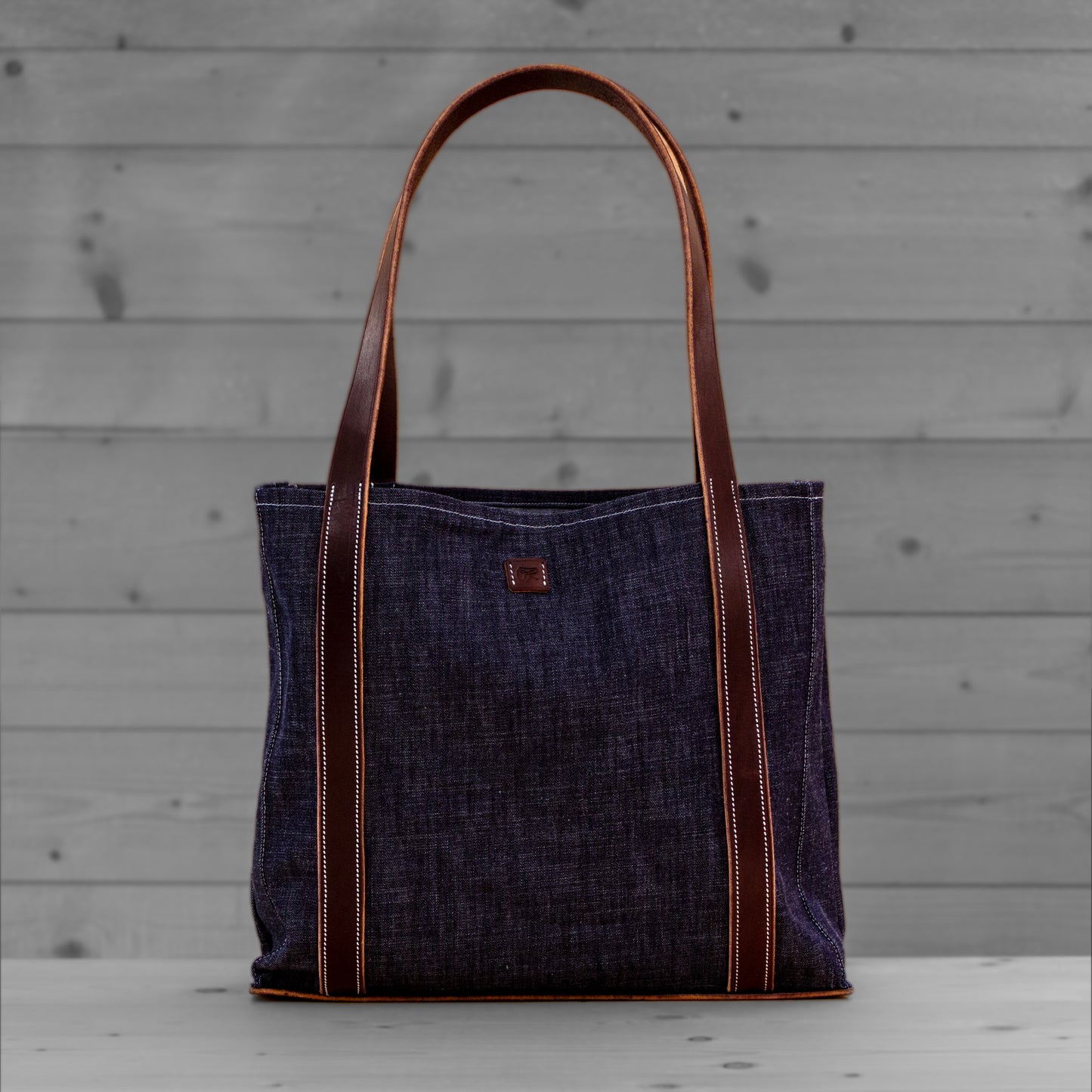 Sac tote en jean avec anses en cuir brun clair, pose sur un fond de bois