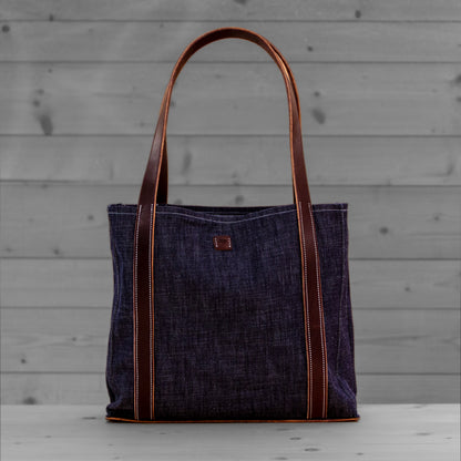 Sac tote en jean avec anses en cuir brun clair, pose sur un fond de bois