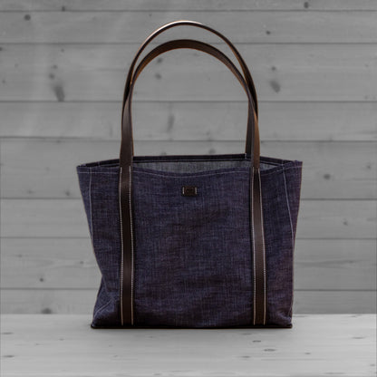 Sac tote en jean avec anses en cuir brun, pose sur un fond de bois